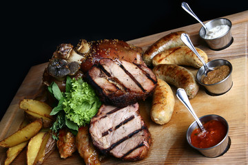 A very large meat dish with steaks, pork knuckle, homemade sausage and baked potatoes. Offer for a male company. Wired board with black background