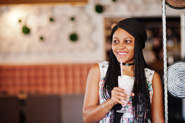 Сheerful african american young woman in summer dress and choker necklace at cafe drinking milkshake cocktail.
