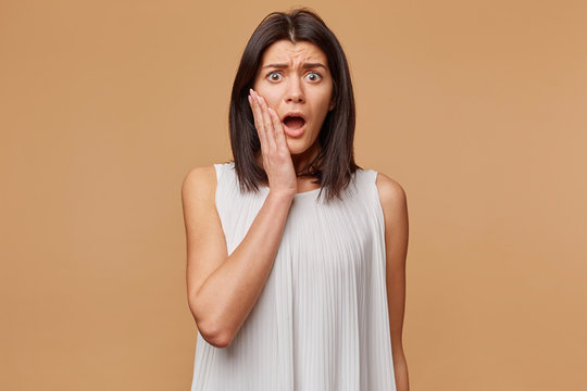 Indoor photo of scared woman in panic nervous frightened, dressed in white dress, keeps hand near cheeck opened mouth as feals fear, isolated on beige background