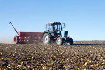 Blue tractor with a red seeder works on the field on a bright sunny autumn day