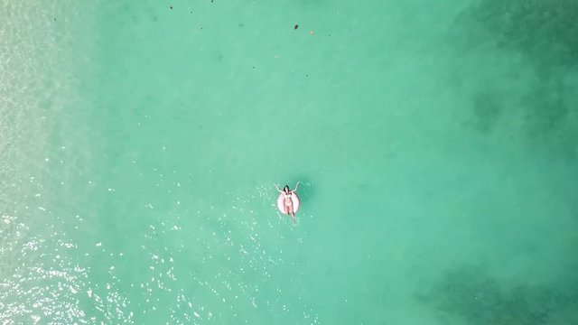 Young woman floating on a donuts buoy floater on a beach with crystal clear water, white sand, boats. 4K drone footage top view.