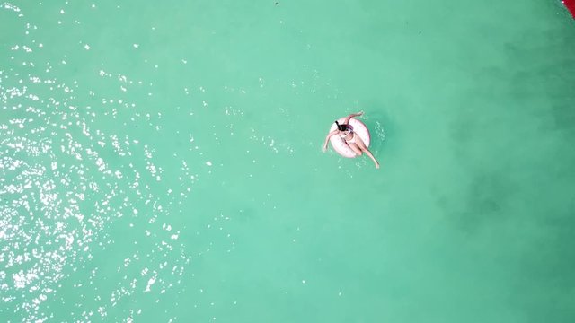 Young woman floating on a donuts buoy floater on a beach with crystal clear water, white sand, boats. 4K drone footage top view.