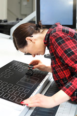  Kobieta drukarz przy stole montażowym ogląda przez lupę jakość wydruku. - obrazy, fototapety, plakaty