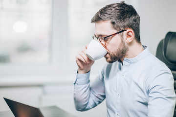 Bearded man office businessman drinking coffee from a cup in the morning at the workplace with a laptop.