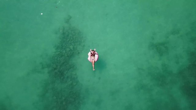 Young woman floating on a donut buoy floater on a beach with crystal clear water, white sand, boats. 4K drone footage top view.