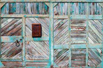 Closed rustic imperfect green garage gate with turquoise peeling paint close-up. Texture of locked wooden metal door with rusty postal box. Flaky dye on grungy wood planks. Rough faded gates of house.