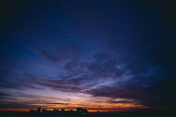 Cityscape with vivid warm dawn. Amazing dramatic blue violet cloudy sky above dark silhouettes of...