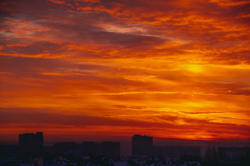 Cityscape with vivid fiery dawn. Amazing warm dramatic cloudy sky above dark silhouettes of city buildings. Orange sunlight. Atmospheric background of sunrise in overcast weather. Copy space.