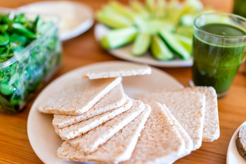 Wooden table setting of healthy vegan vegetarian lunch with macro closeup of plain white rice cakes galettes squares and lettuce green vegetables with bokeh background and juice