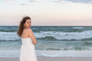 Fototapeta na wymiar Young woman standing in white dress on beach evening in Florida panhandle shivering in wind by ocean waves looking back sad