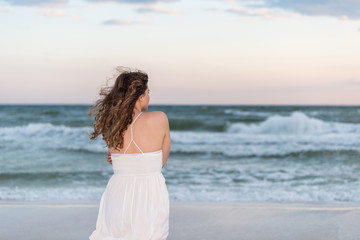 Fototapeta na wymiar Young woman back standing in white dress on beach evening in Florida panhandle shivering arms crossed in wind by ocean waves