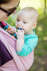 Young woman carrying her baby daughter in woven wrap outdoors in spring park. Baby girl chewing teething beads.