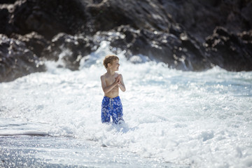 Young boy looking at big waves while standing in shallow water in rough sea