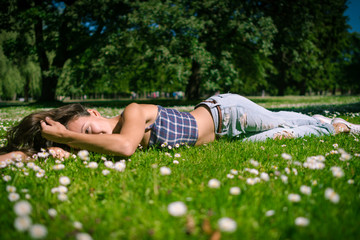 Naklejka na ściany i meble Young joyful woman lies on green grass. Happy girl is relaxing at chamomile field in warm summer day on background of big trees in park.