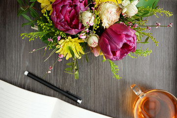 Bright bouquet of flowers with notepad and tea on a wooden table, top view