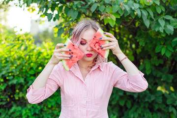 Fototapeta premium Beautiful young woman with pink hair holding two slices of watermelon in front of her face