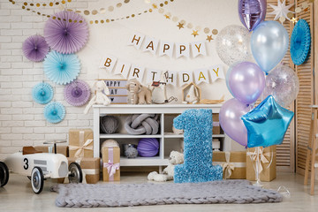 Birthday blue and purple decorations with gifts, toys, garlands and figure for little baby party on a white bricks background.