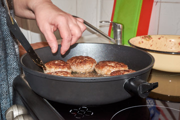 A woman frying meat patties in a pan turns them over with a spatula and helps herself with her hands.