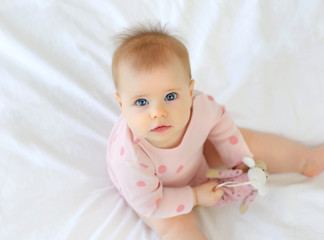 Charming blue-eyed baby with a funny hairstyle 9 months sitting in bed in pink clothes. Top view