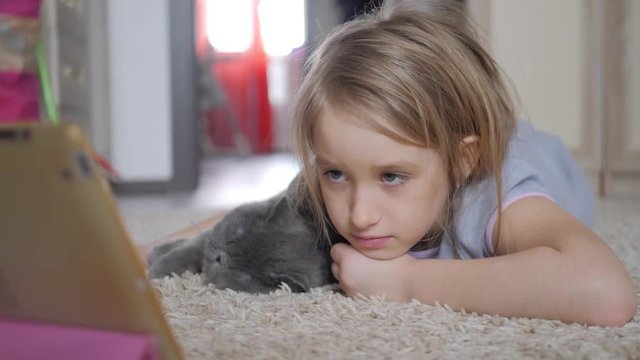 Cute little girl holding digital tablet and hugging your favorite cat lying on the floor. Beautiful girl playing on tablet pc. Education, school, technology and internet concept.