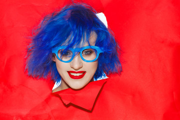 Portrait of an extravagant woman with blue hair and glasses looking through hole in paper wall