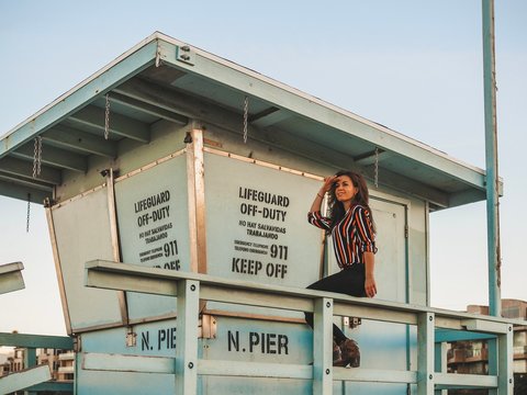 Brunette girl in striped bright shirt with long hair sits on lifeguard tower near Manhattan beach pier in Los Angeles at sunrise