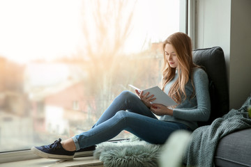 Beautiful young woman reading book while sitting on window sill - Powered by Adobe