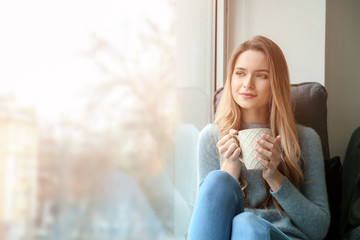 Beautiful young woman drinking coffee while sitting on window sill
