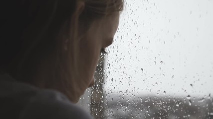 Sad teenager girl looking outside through the window in the rainy day.