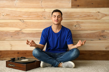 Fototapeta premium Young man meditating and listening to music on record player near wooden wall