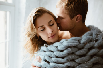 Stylish interior. A couple near the window, under one blue blanket of large mating. Gentle embrace and kisses. Blonde. Floor lamp. Athletic build. Gray pajamas in hearts. Love story.