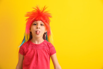 Funny little girl in wig showing tongue on color background. April fools' day celebration