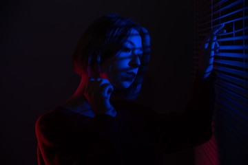 A beautiful girl with a chain in her hands stands near the window with blinds in the light of neon lights of blue and red light