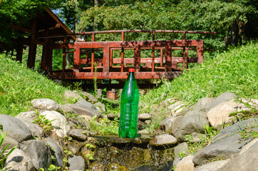 Green plastic bottle of water in a stream with a small wooden bridge behind