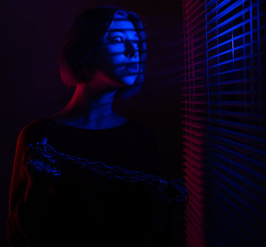 A beautiful girl with a chain in her hands stands near the window with blinds in the light of neon lights of blue and red light