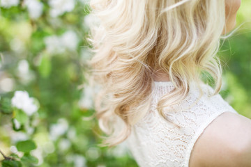 Fashion photo of beautiful young woman  posing in garden with blossom trees. Blonde in flowering gardens. Beautiful streaming blonde curls.  View from a back. Close up picture