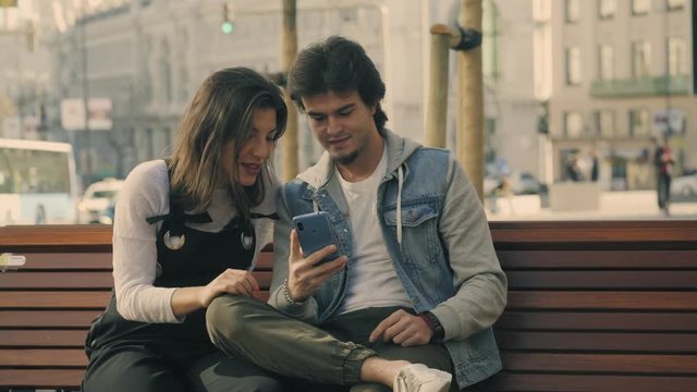 Pareja de j&oacute;venes sentados en un banco en la calle de una ciudad mirando un smartphone