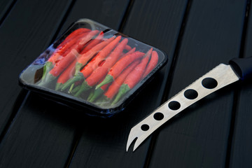 A knife for cheese and vegetables, along with a package of red chili peppers, lie on a black wooden surface