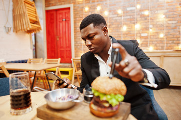  Respectable young african american man in black suit sitting in restaurant with tasty double burger and soda drink. Looking at phone before eat.