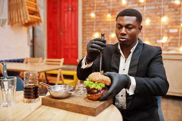Respectable young african american man in black suit and gloves for food sitting in restaurant with tasty double burger and soda drink.