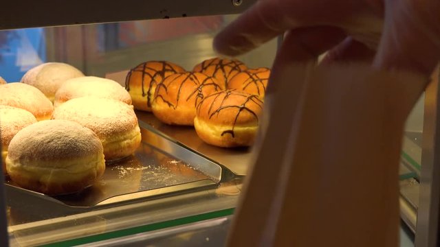 A clerk takes a Berliner doughnut off a display shelf on a counter and puts it into a disposable paper bag - closeup