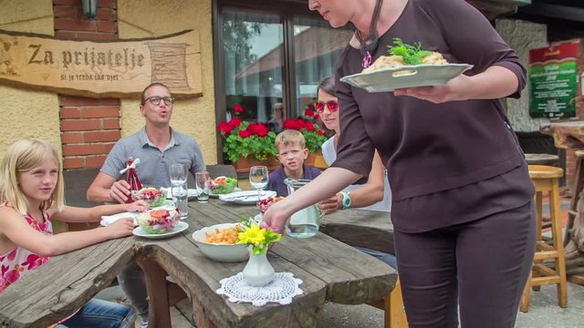 Young family is sitting outside in a restaurant. A waitress bring French fries and mashed potatoes and puts them on a table.