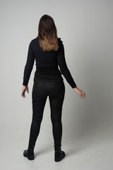 full length portrait of a brunette girl wearing  modern black jacket and pants, standing pose with back to the camera on grey studio background. © faestock
