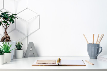 Hipster desk at white empty wall with space for text. Copy space. Open notebooks and wooden pencils in a gray mug. Green succulents, bonasi and the letter A as decoration.