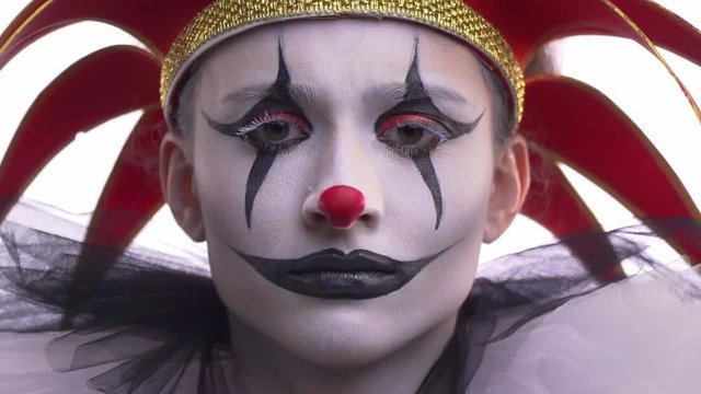 Close up of the jester with makeup and a red hat with little bells, twitching eyebrows