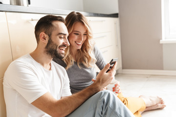 Amazing young loving couple sitting at the kitchen using mobile phone on a floor.