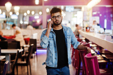 Stylish asian man wear on jeans jacket and glasses posed against bar in club and speaking on mobile phone.