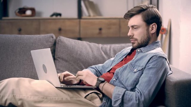 Freelancer in denim shirt lying on couch with laptop, typing on keyboard and blinking while looking at screen