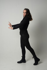 full length portrait of a brunette girl wearing  modern black jacket and pants, standing pose with back to the camera on grey studio background.