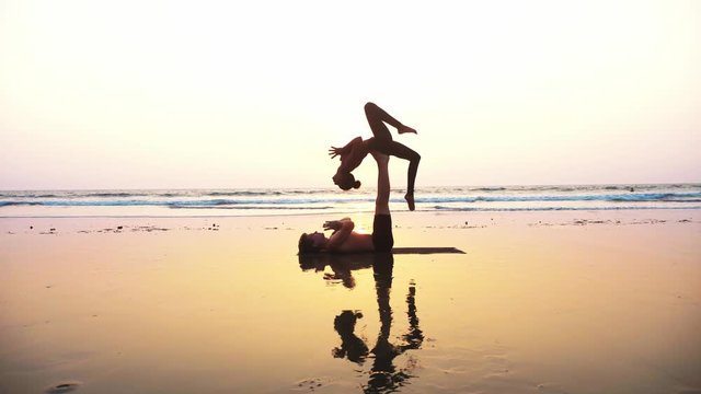 Silhouette of fit sporty couple practicing acrobatic yoga with partner together on the beach in slow motion. Steadicam shot of female acrobat balancing on feet of her partner in fylfot shape